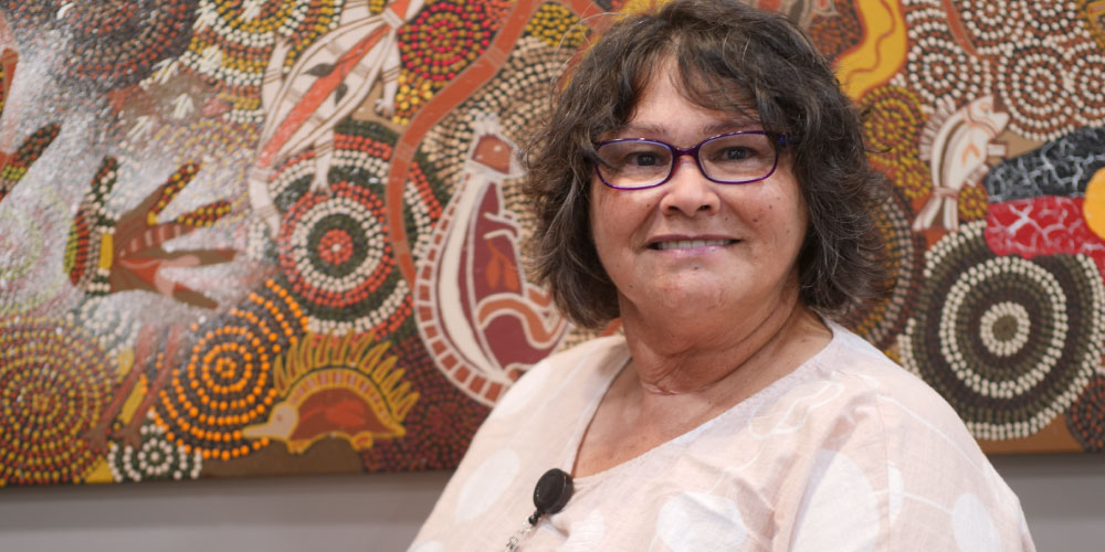 Aunty Gina Bundle faces the camera smiling. She is standing in front of a brown, red, yellow and white Aboriginal dot painting.