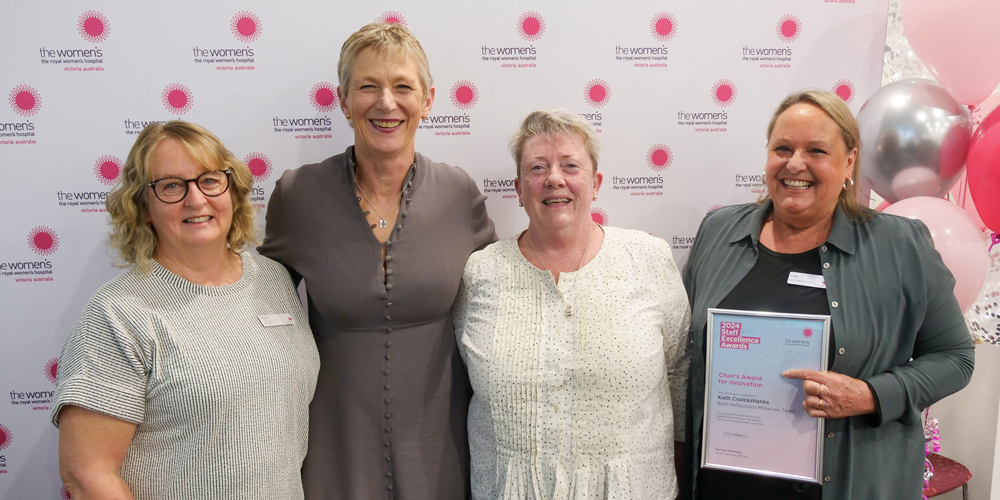 A group of four smiling people in front of the Women's signage and balloons. One is holding a Staff Excellence Awards certificate.