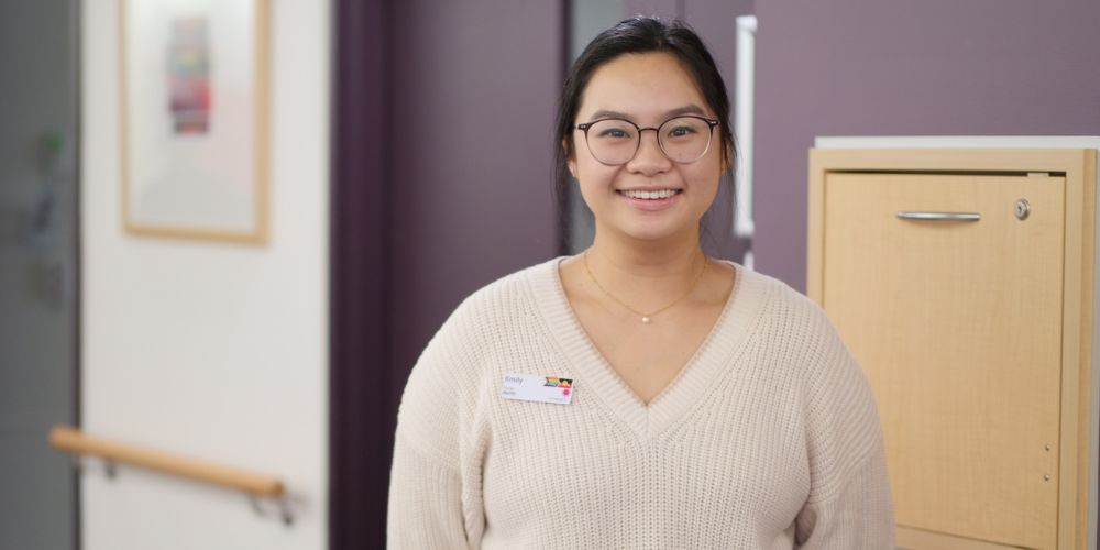 Nurse Emily Jong stares in to camera smiling, standing in a hallway in a hospital ward.