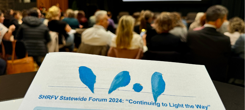 Program of SHRFV forum 2024 held up in front. Background shows rows of forum attendees. 