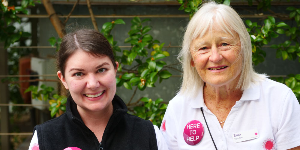 Two volunteers smiling at the camera against a green backdrop