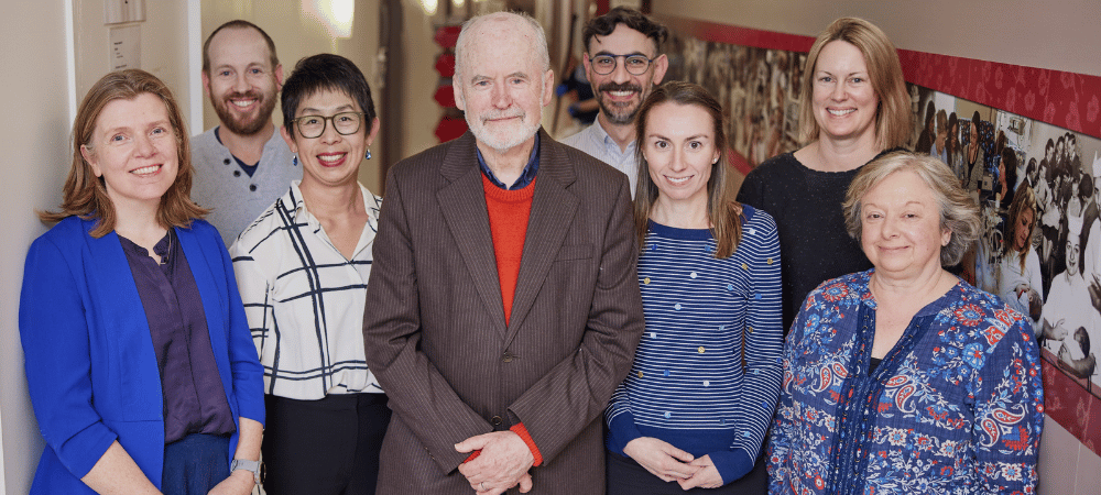 Seven researchers (five of whom are women) are smiling as they stand with an older man, Professor Lex Doyle.