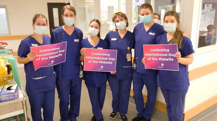Six midwives in scrubs and surgical masks holding signs saying 